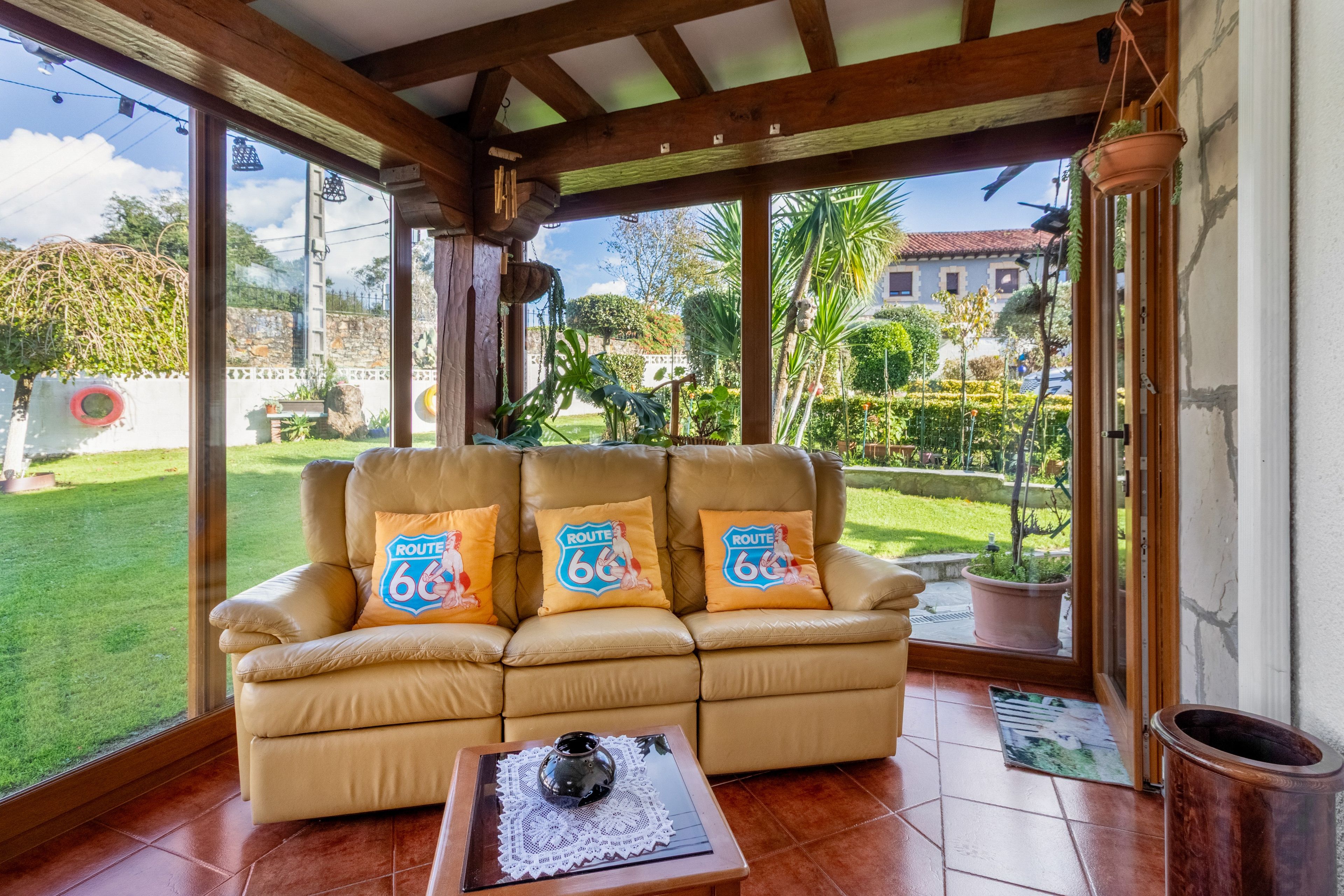 A tan leather sofa with Route 66 pillows, a small table with a teapot, and a view of a garden through large windows.