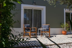A patio with two wooden chairs and a small table, a potted plant, and a window with a view. - Farm Lodges Olive, Casa Alfarrobeira, Moncarapacho Countryside (moncarapacho)