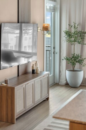 Ein modernes Wohnzimmer mit einem Wand-Fernseher, einem Holzschrank mit Dekorationsobjekten, einer großen Topfpflanze und einer Glastür, die zu einem Außenbereich führt.