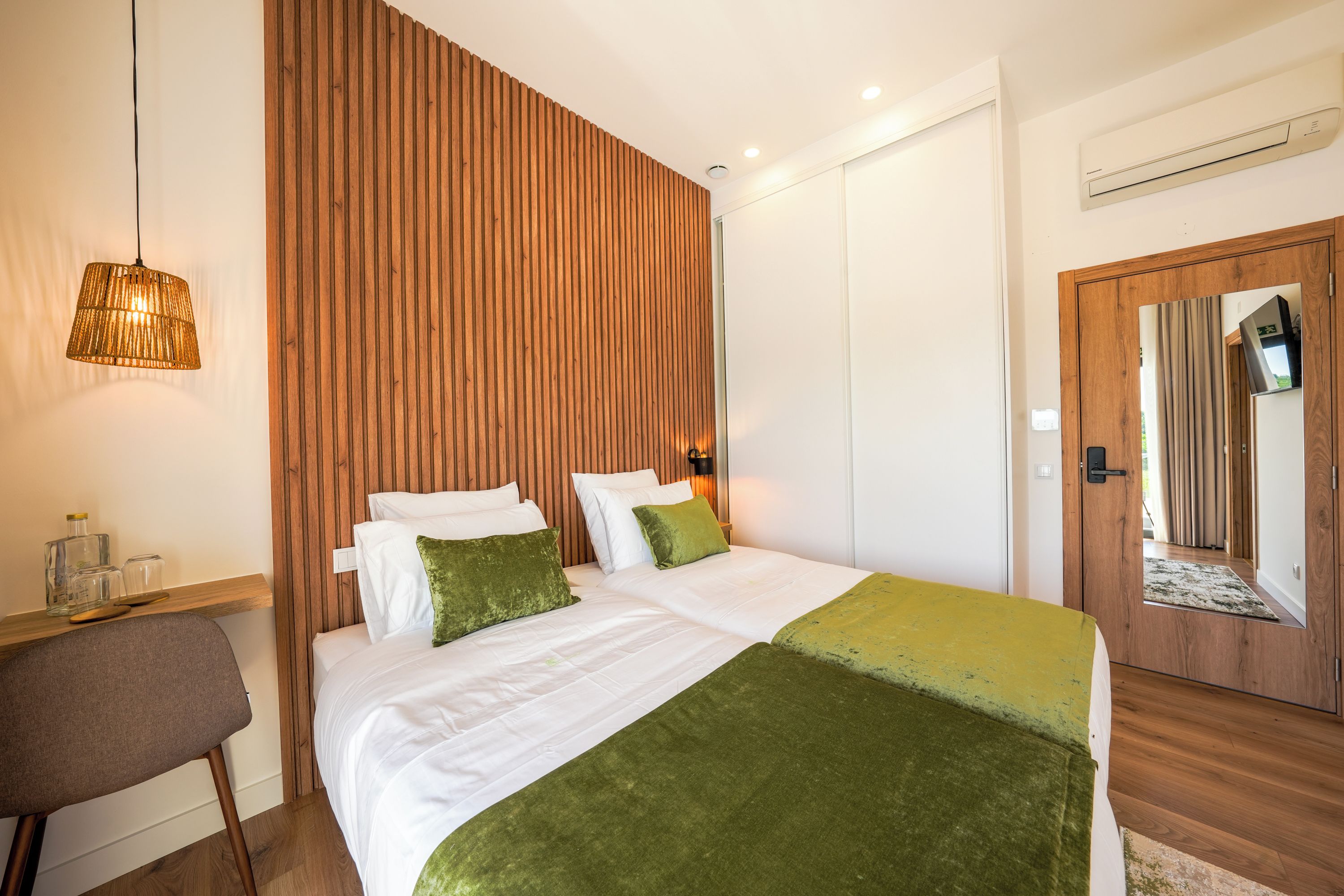Un dormitorio con pared de listones de madera, una cama con ropa blanca y almohadas verdes, una silla de madera, una lámpara colgante y una puerta con espejo.