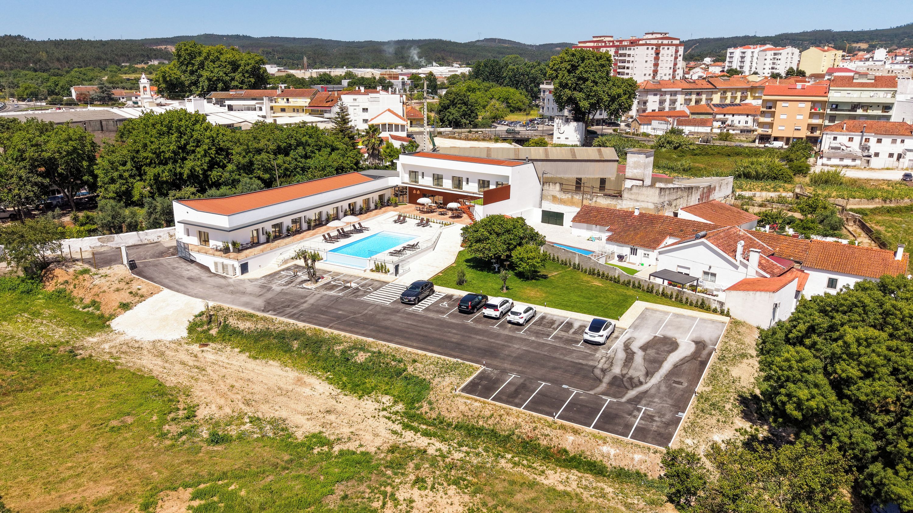 An aerial view of a hotel with a swimming pool, parking lot, and surrounding greenery.