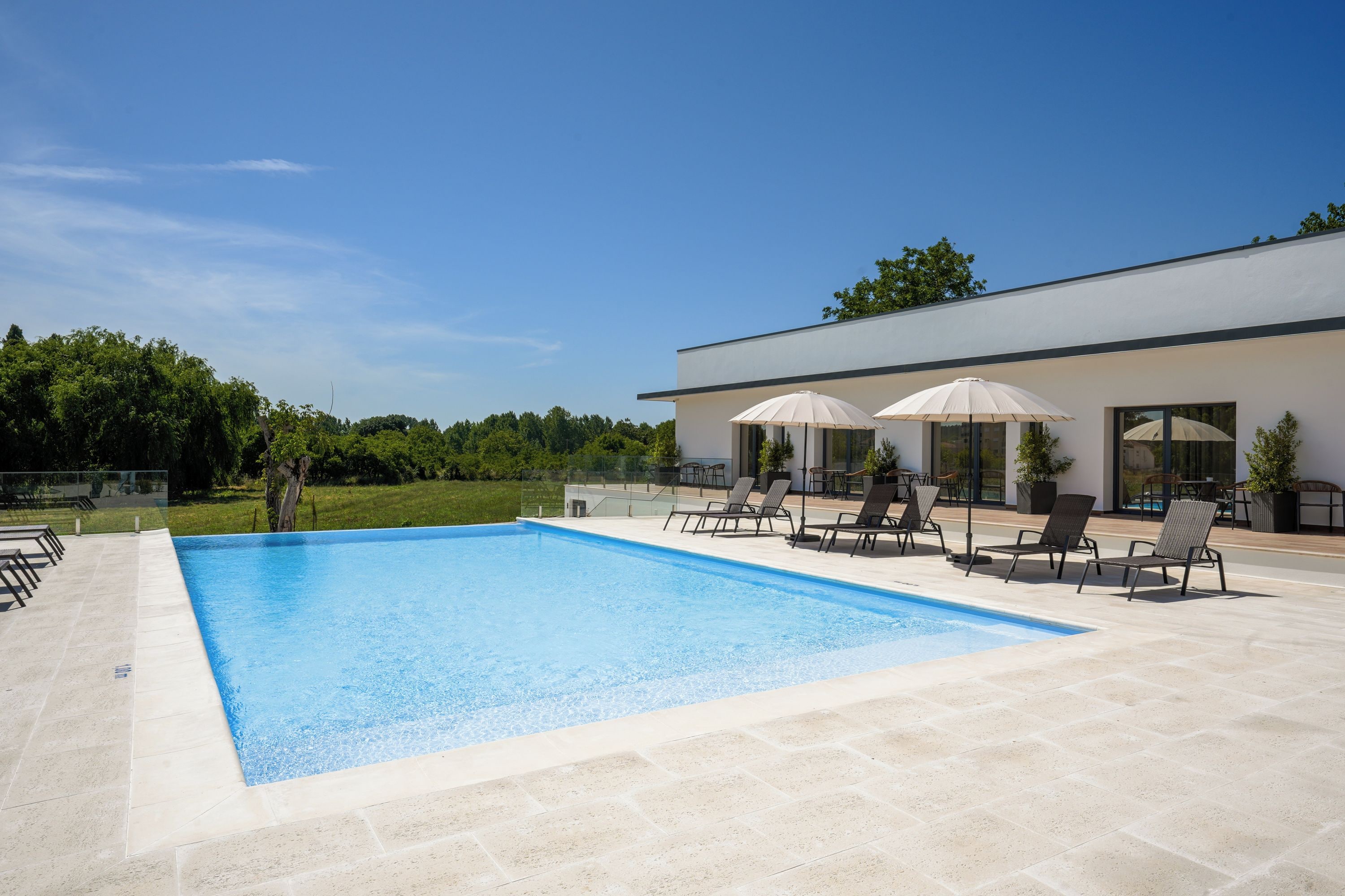 Une piscine entourée de chaises longues et de parasols, elle-même bordée d’un bâtiment moderne doté de grandes fenêtres.