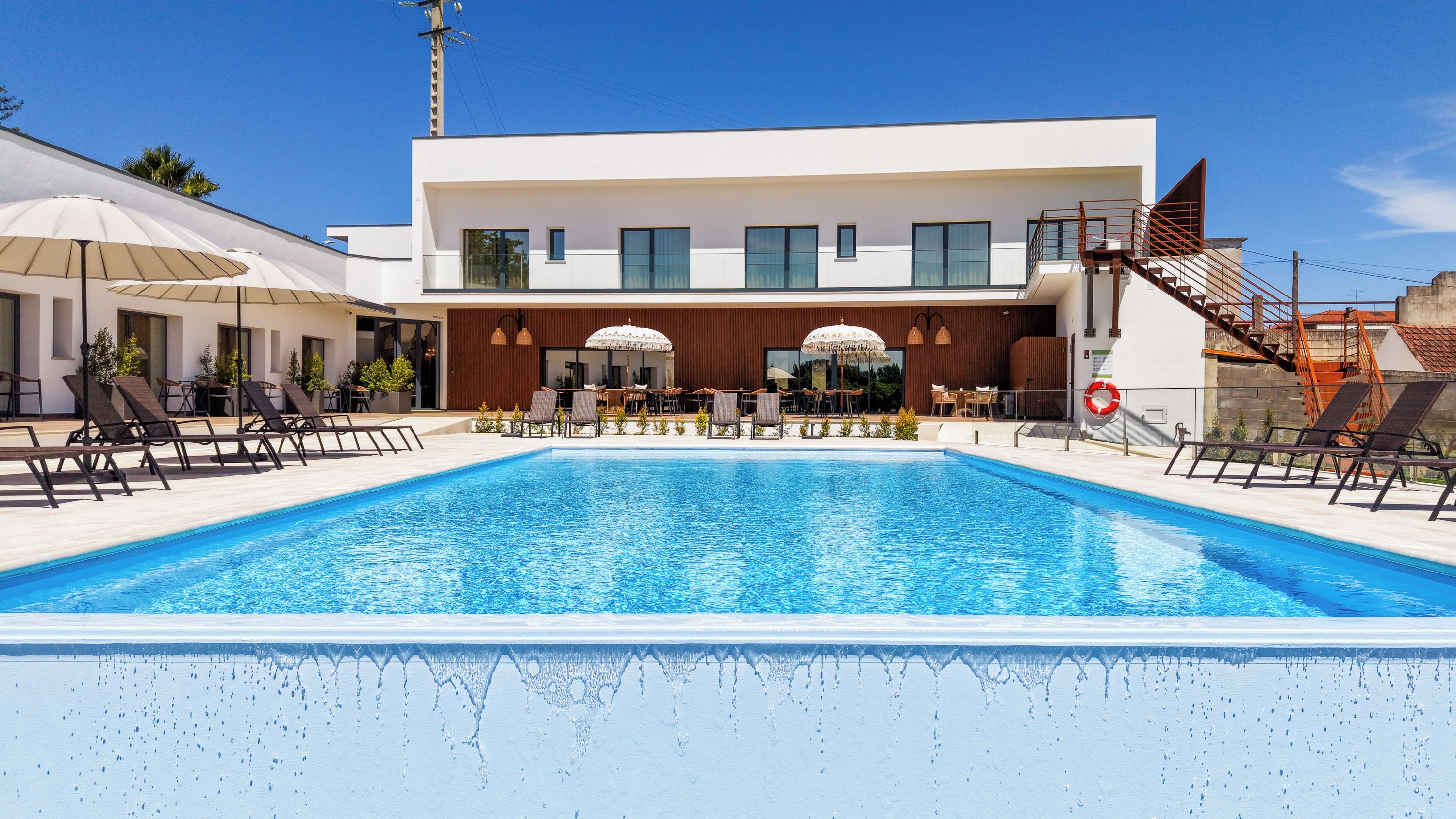 Un área de piscina moderna con sillones de descanso y sombrillas, una piscina azul cristalina y un edificio contemporáneo con grandes ventanales.