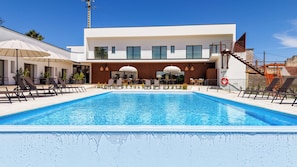 Un área de piscina moderna con sillones de descanso y sombrillas, una piscina azul cristalina y un edificio contemporáneo con grandes ventanales.