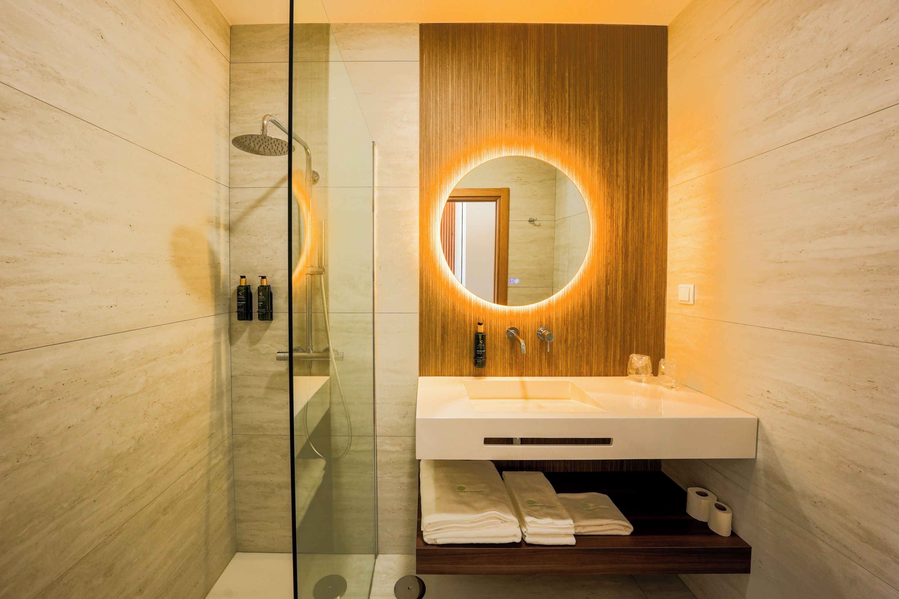 A modern bathroom with a glass-enclosed shower, a wooden vanity with a built-in sink, and a mirror with a circular light.
