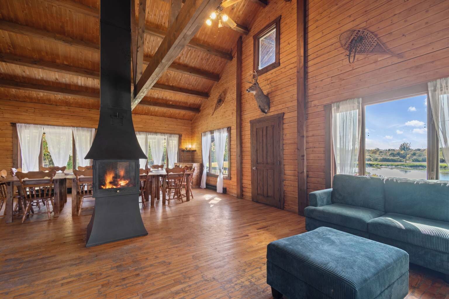 Interior de una cabaña de troncos con chimenea, muebles de madera y ventanas amplias que ofrecen vistas al exterior.