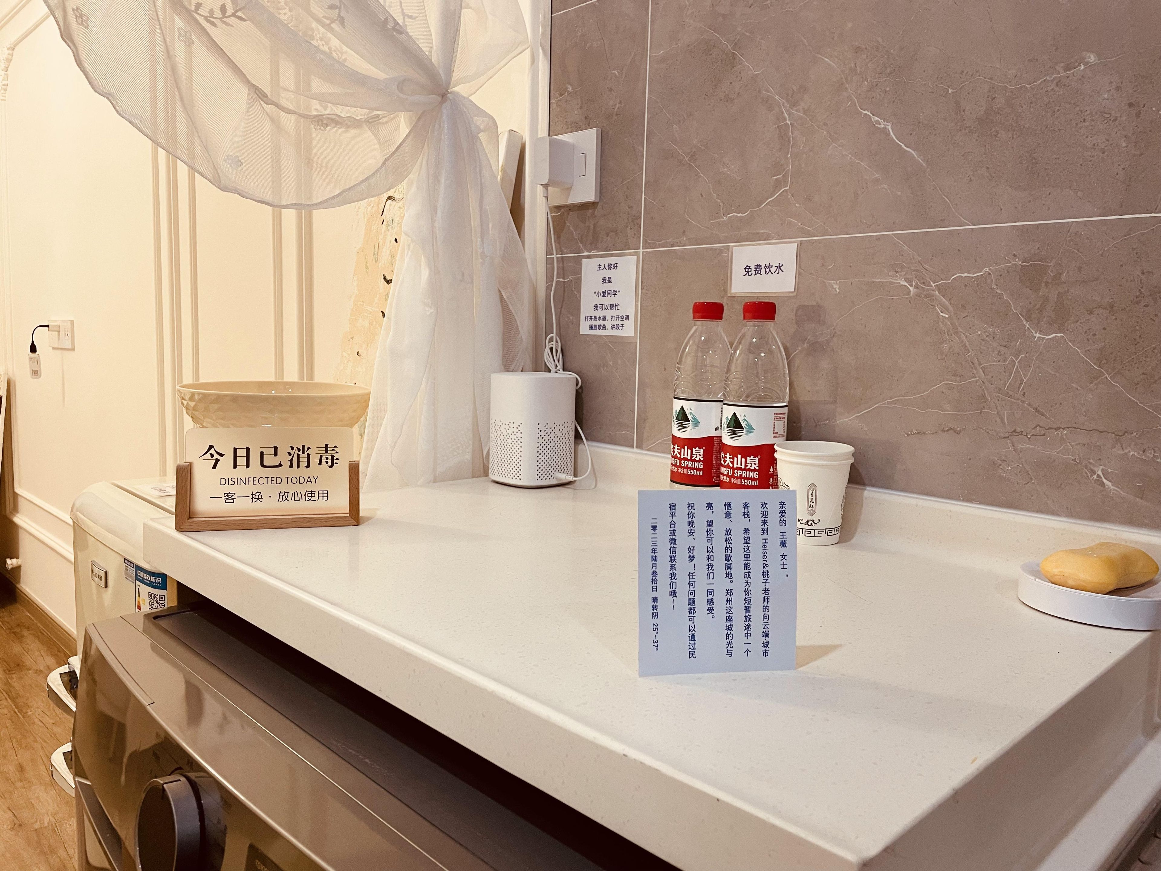 Mostrador de hotel con un lavabo blanco, una mosquitera, una taza blanca, dos botellas de agua y un cartel que indica agua gratuita.