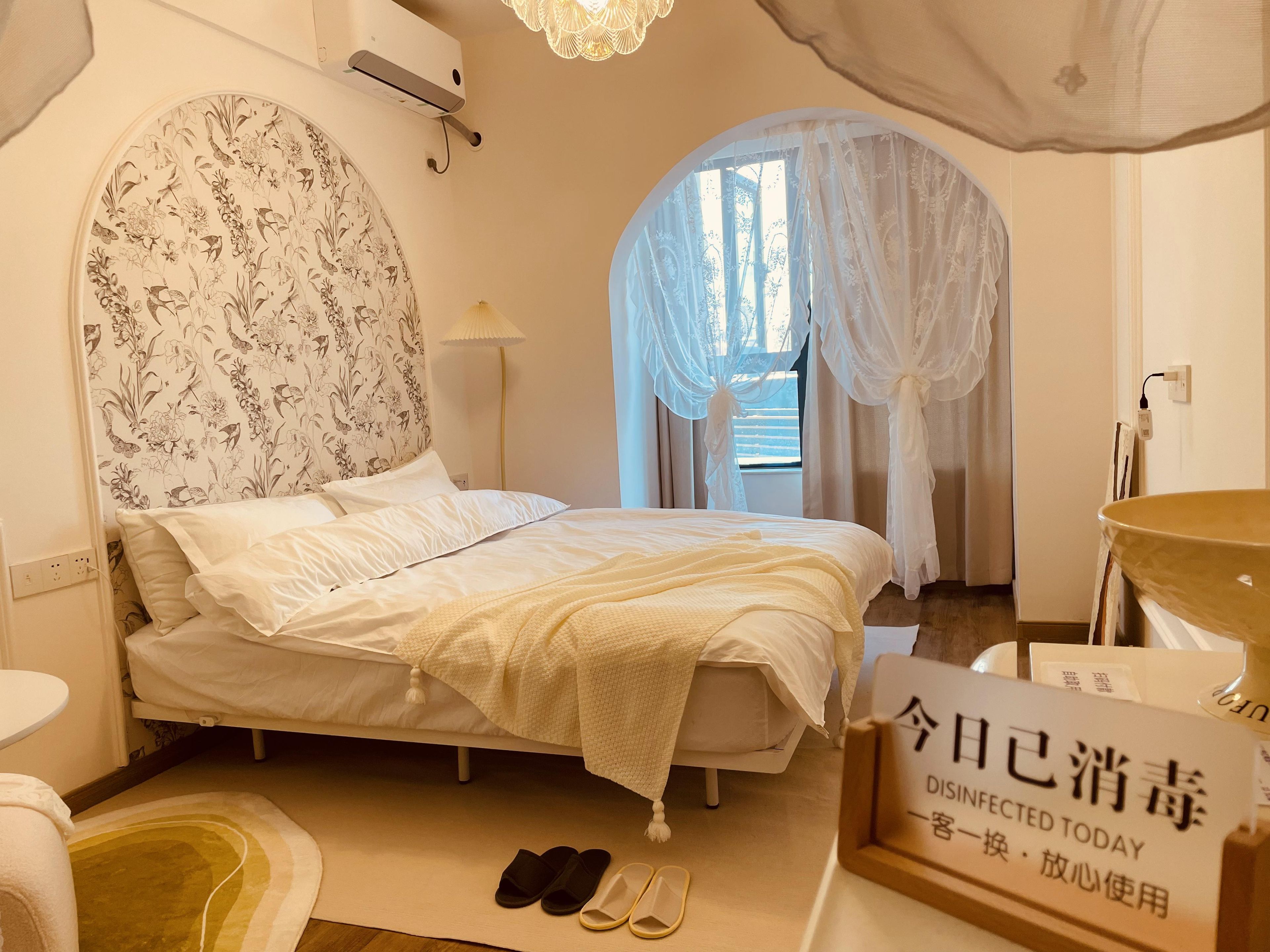 A hotel room with a bed, a bedside table, a lamp, a sign indicating the room is disinfected, and a window with sheer curtains.