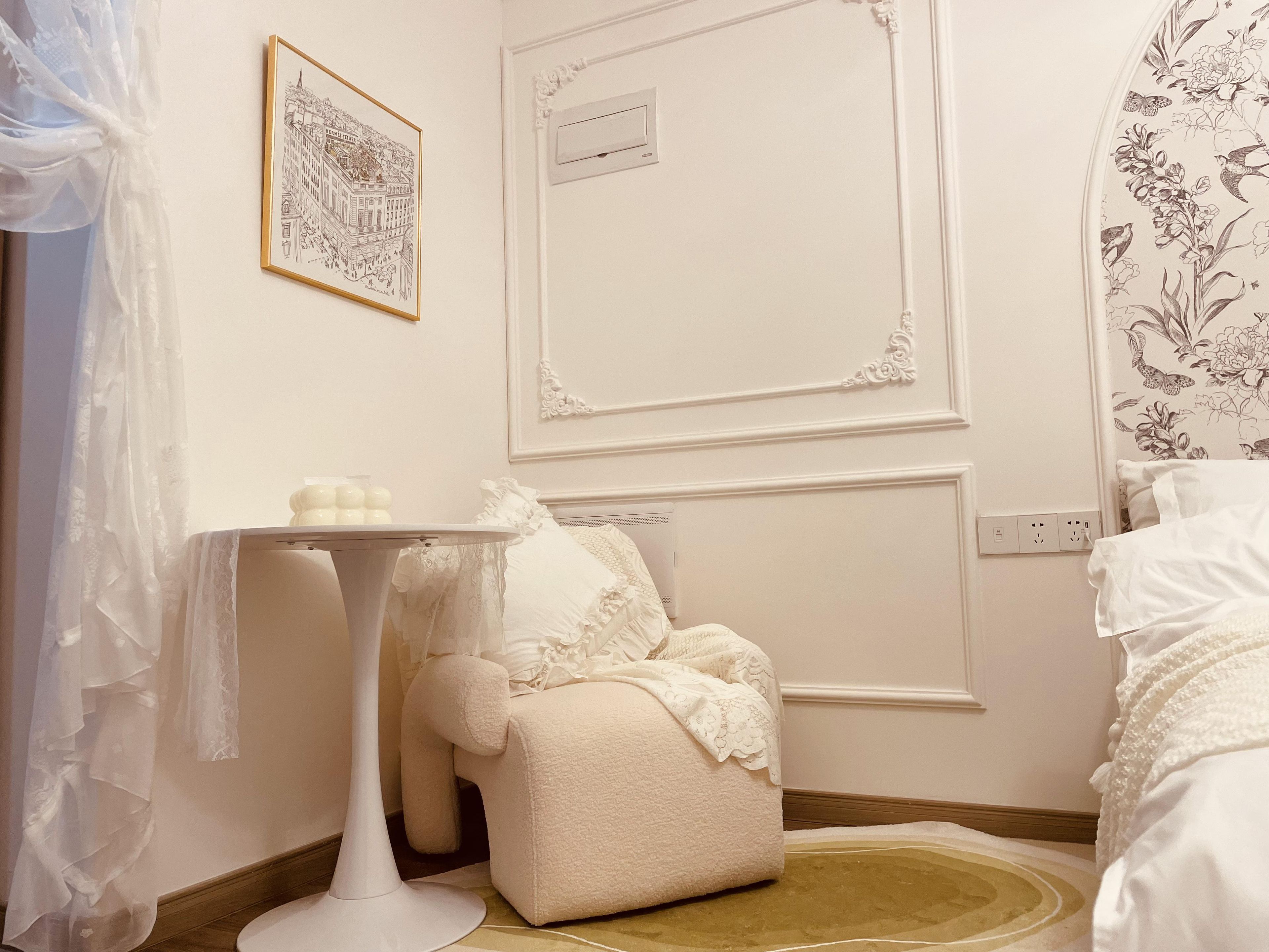Una habitación con cama blanca, una mesita de noche con lámpara, un cuadro en la pared y un estampado floral en el papel tapiz.