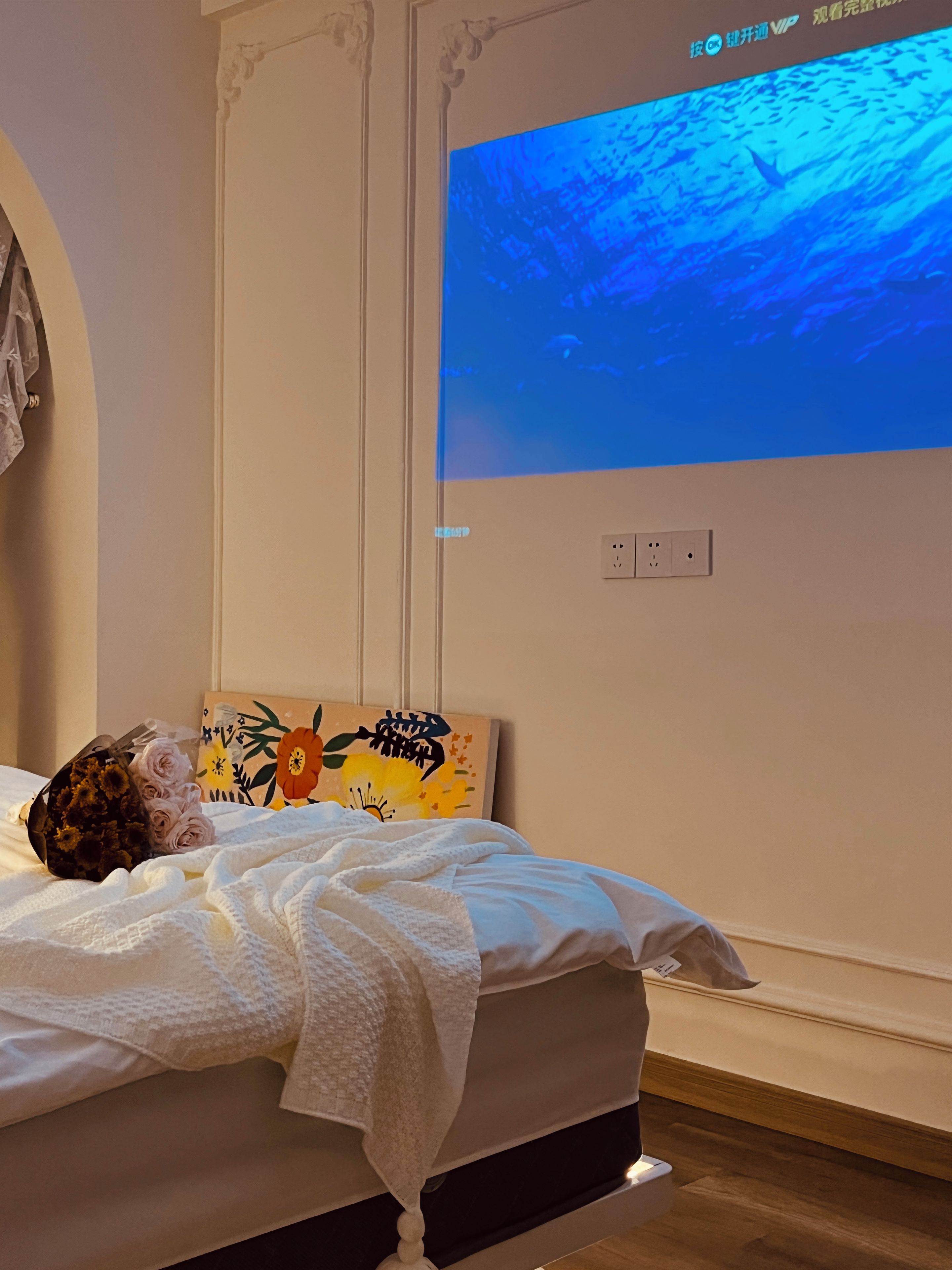 A bed with a white blanket, a bouquet of flowers, and a wall-mounted TV displaying a blue underwater scene.