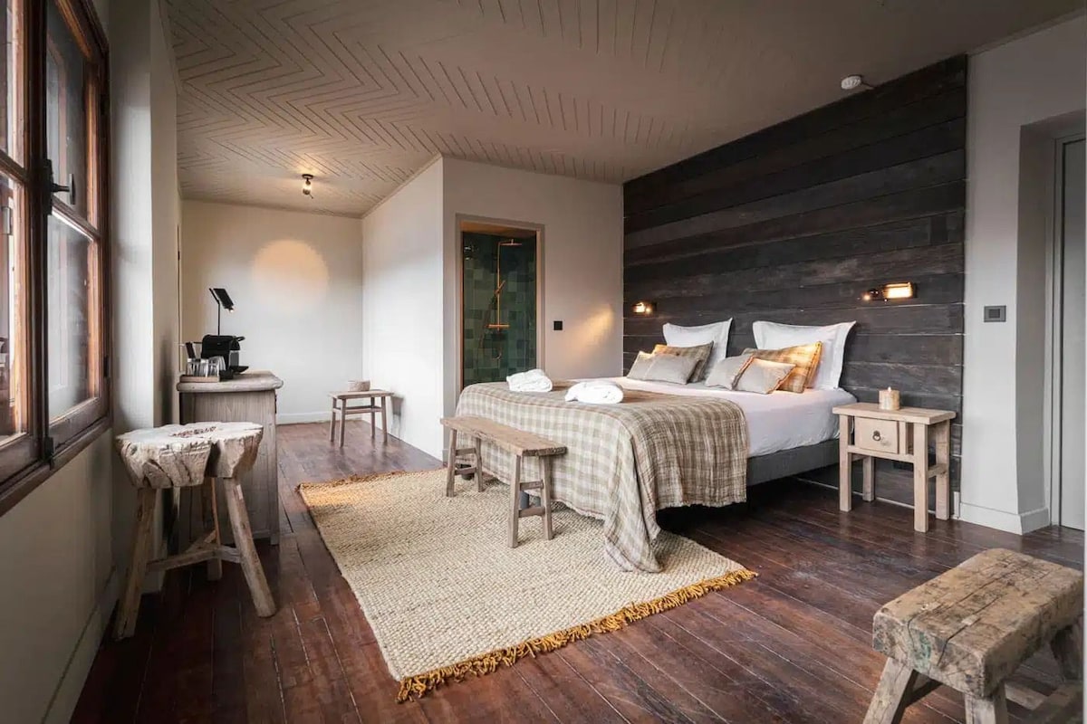 A bedroom with a large bed, bedside tables, a bench, a small stool, and a woven rug.