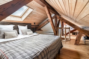 A loft bedroom with a sloped ceiling, wooden beams, a skylight, a bed with a patterned bedspread, and a bedside table. - Le Refuge du Grand Hôtel (Chantemerle)