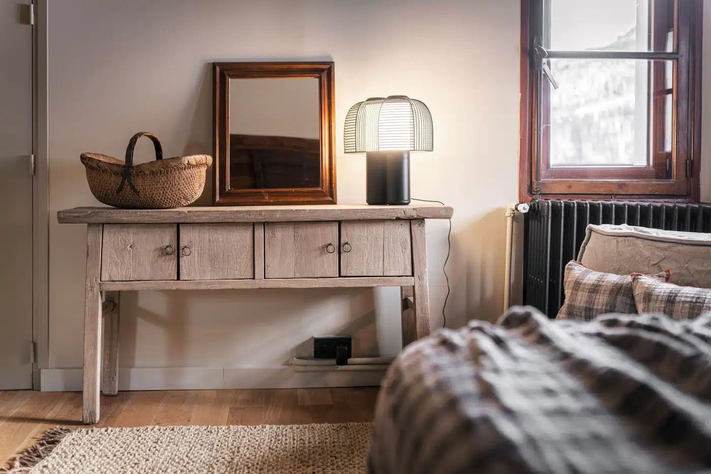 Een slaapkamer met een houten dressoir, een spiegel, een lamp, een rieten mand en een bed met een geruit plaid.