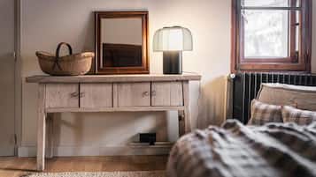 Een slaapkamer met een houten dressoir, een spiegel, een lamp, een rieten mand en een bed met een geruit plaid.