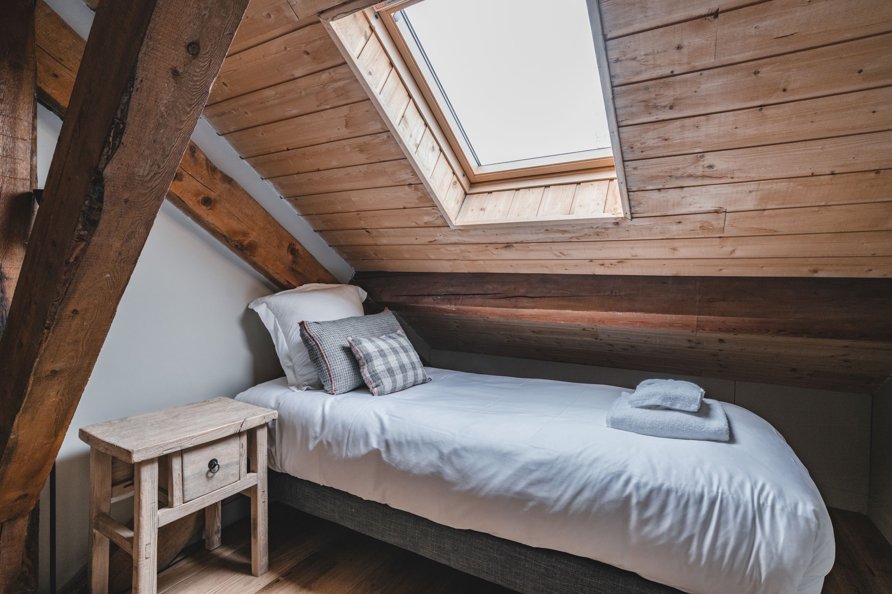 Une petite chambre à coucher accueillante, avec un plafond en pente et un puits de lumière, comprenant un lit simple avec des oreillers et une table de chevet.