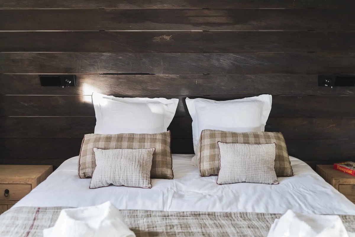 A bed with white linens and two pillows, a wooden headboard, and a bedside table with a lamp.