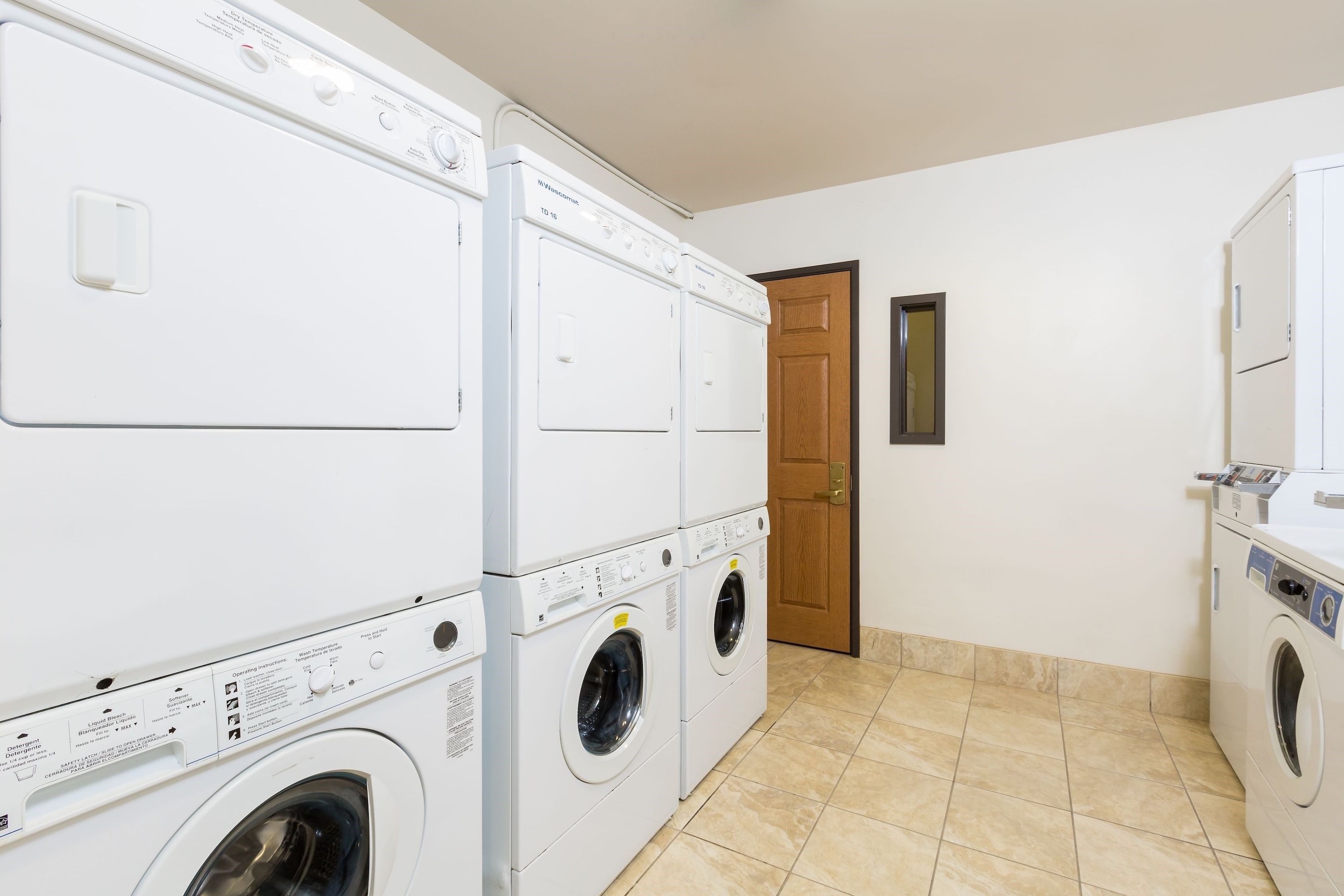 Une buanderie avec des machines à laver et des sèche-linge superposés, une porte en bois et un sol carrelé.