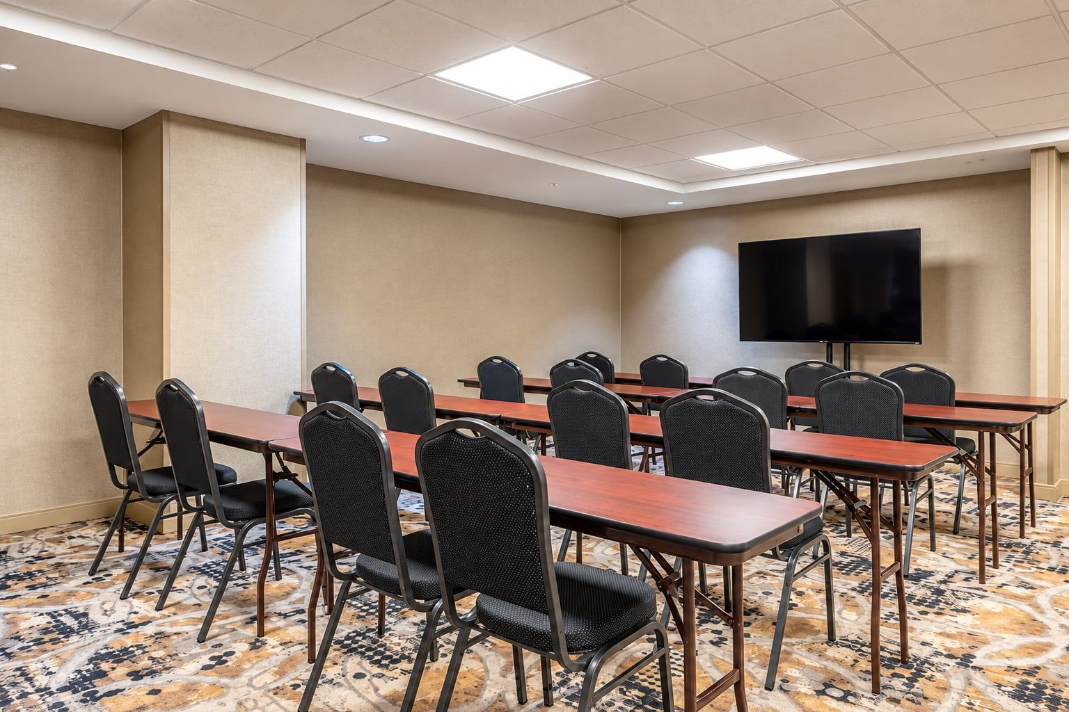 Une salle de conférence équipée de plusieurs tables et chaises, d’un téléviseur à écran plat et d’un revêtement de sol en moquette à motifs.