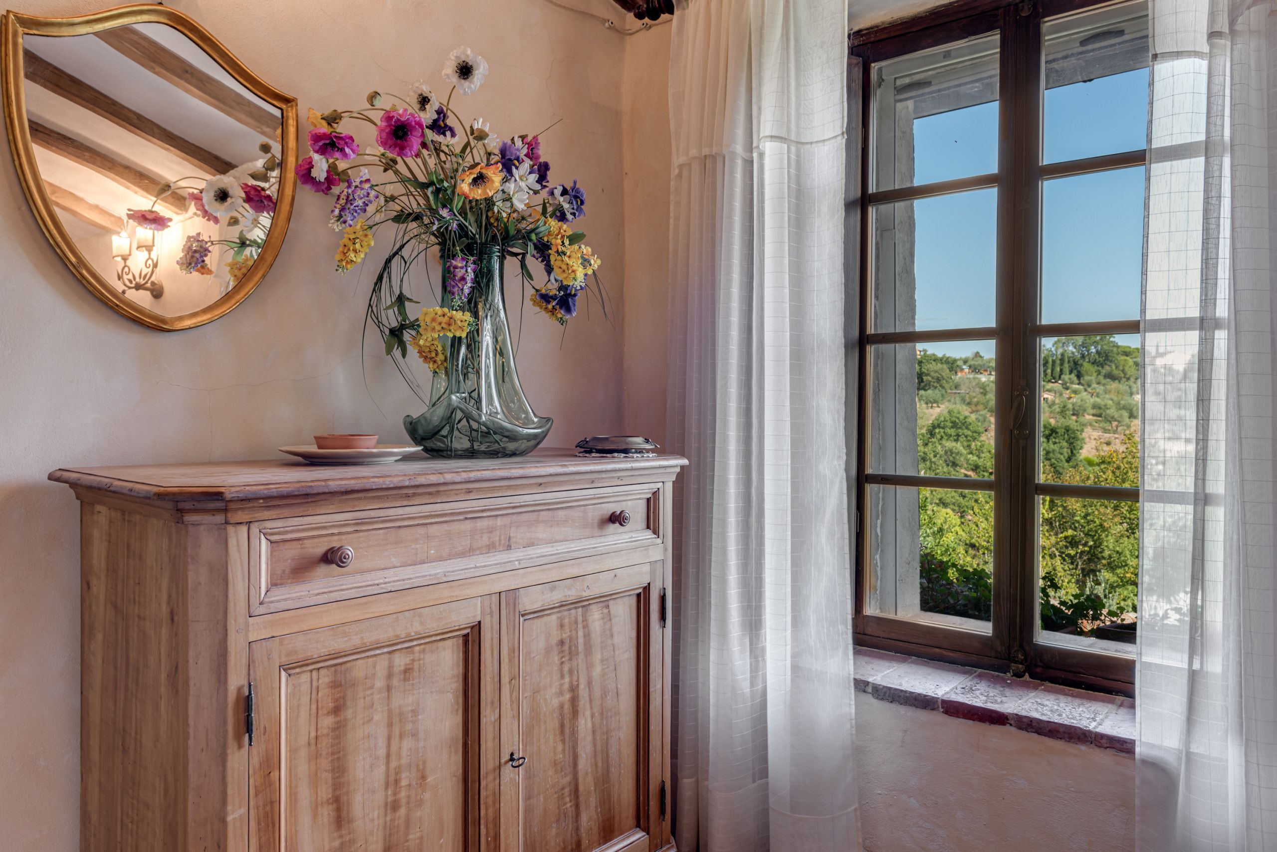 Un meuble en bois orné d’un vase de fleurs, d’un miroir et d’une fenêtre donnant sur un paysage verdoyant.