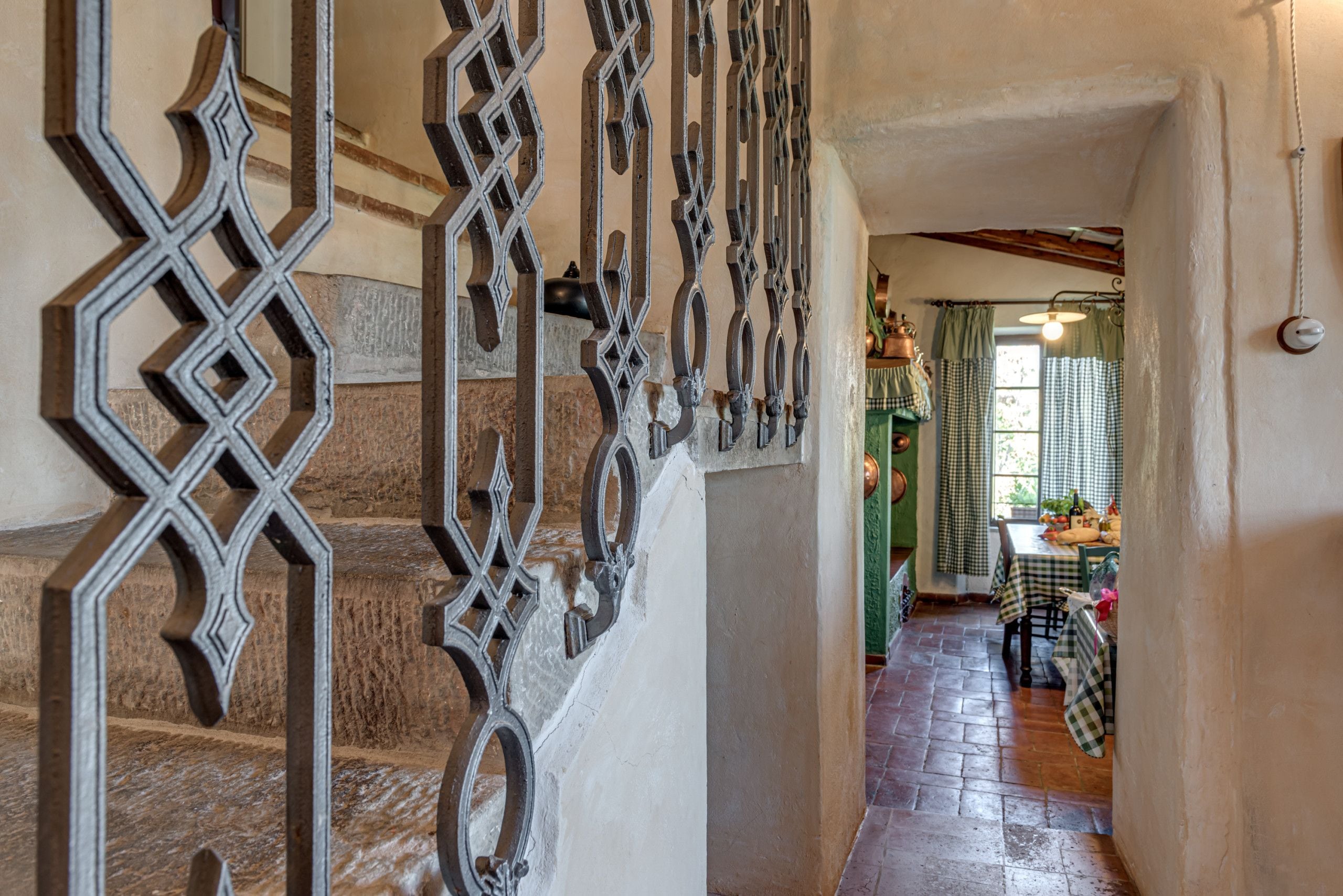 Un escalier orné de rampes en métal menant à une pièce où se trouve une table recouverte d’une nappe à carreaux et un luminaire suspendu.