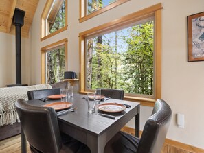 A dining area with a table set for four, large windows with a forest view, and a fireplace. - Riversong Retreat Sandy River View Cabin (Mount Hood Village)