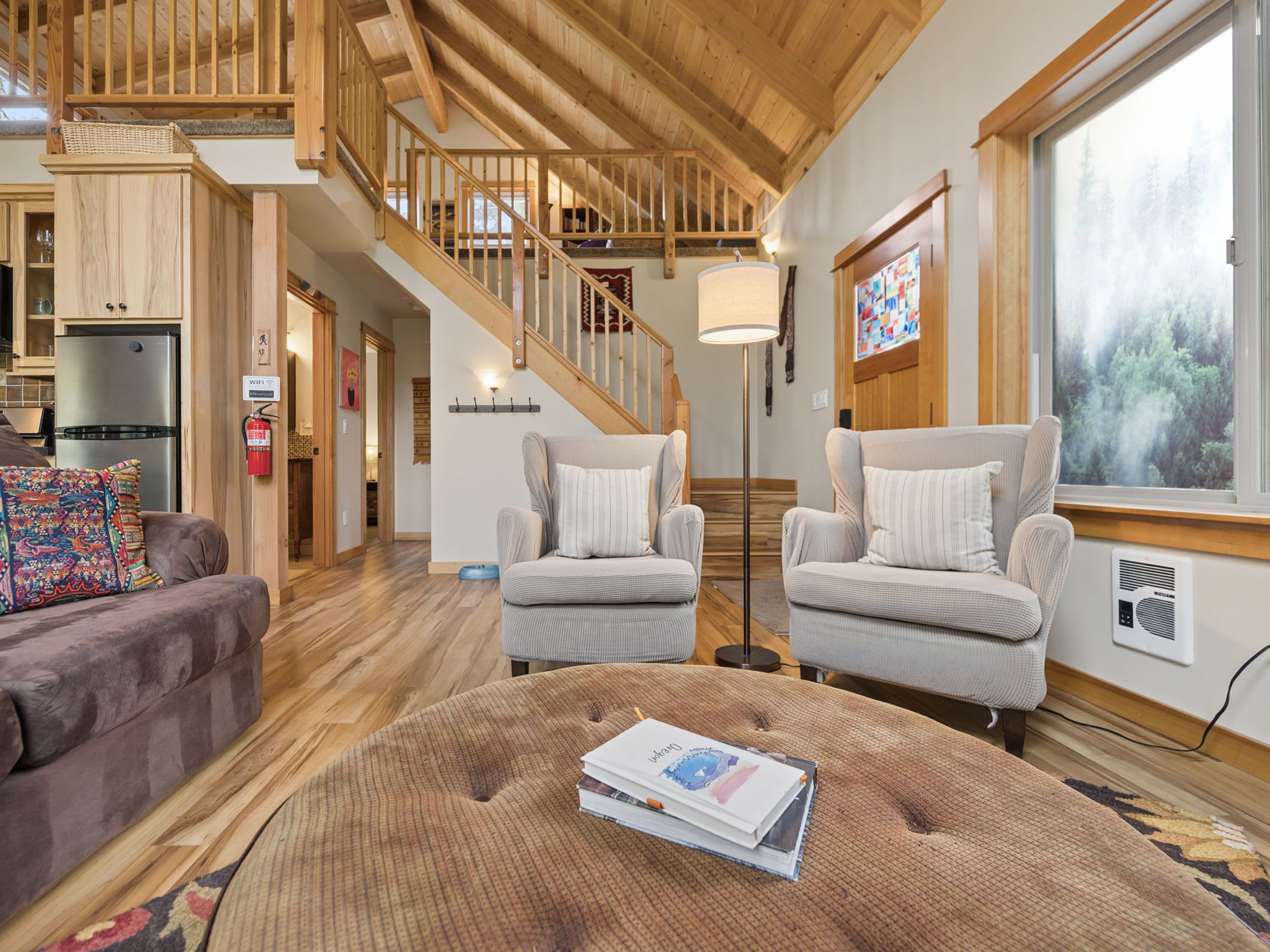 A spacious living area with a wooden staircase, two armchairs, a sofa, and a large window with a forest view.