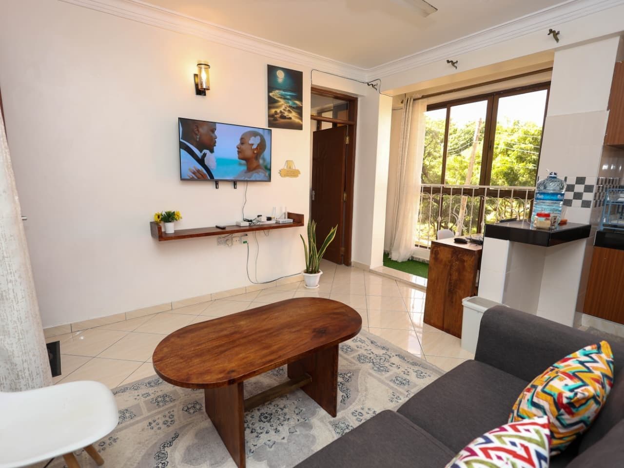 A living room with a TV, sofa, and a small table. There is a balcony with a view of greenery.