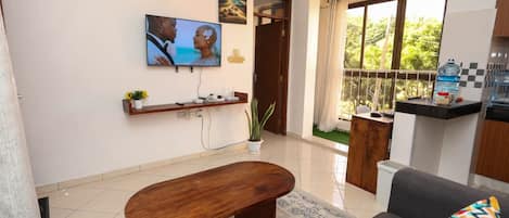 A living room with a TV, sofa, and a small table. There is a balcony with a view of greenery.