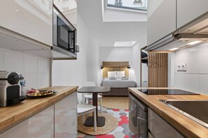 Een compacte hotelkamer met een kitchenette, een bed met hoofdeinde, een klein zithoekje en uitzicht op de stad door een raam.