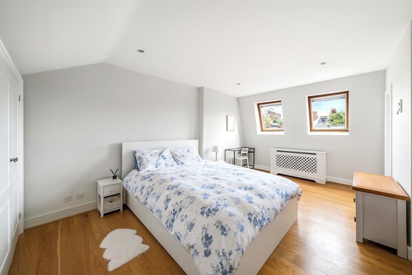 Une chambre à coucher avec un lit, une table de chevet, une chaise, un radiateur, des fenêtres et un tapis blanc.