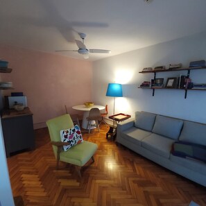 En stue med ventilator, et rundt bord med stole, en blå lampe, en sofa og en bogreol.