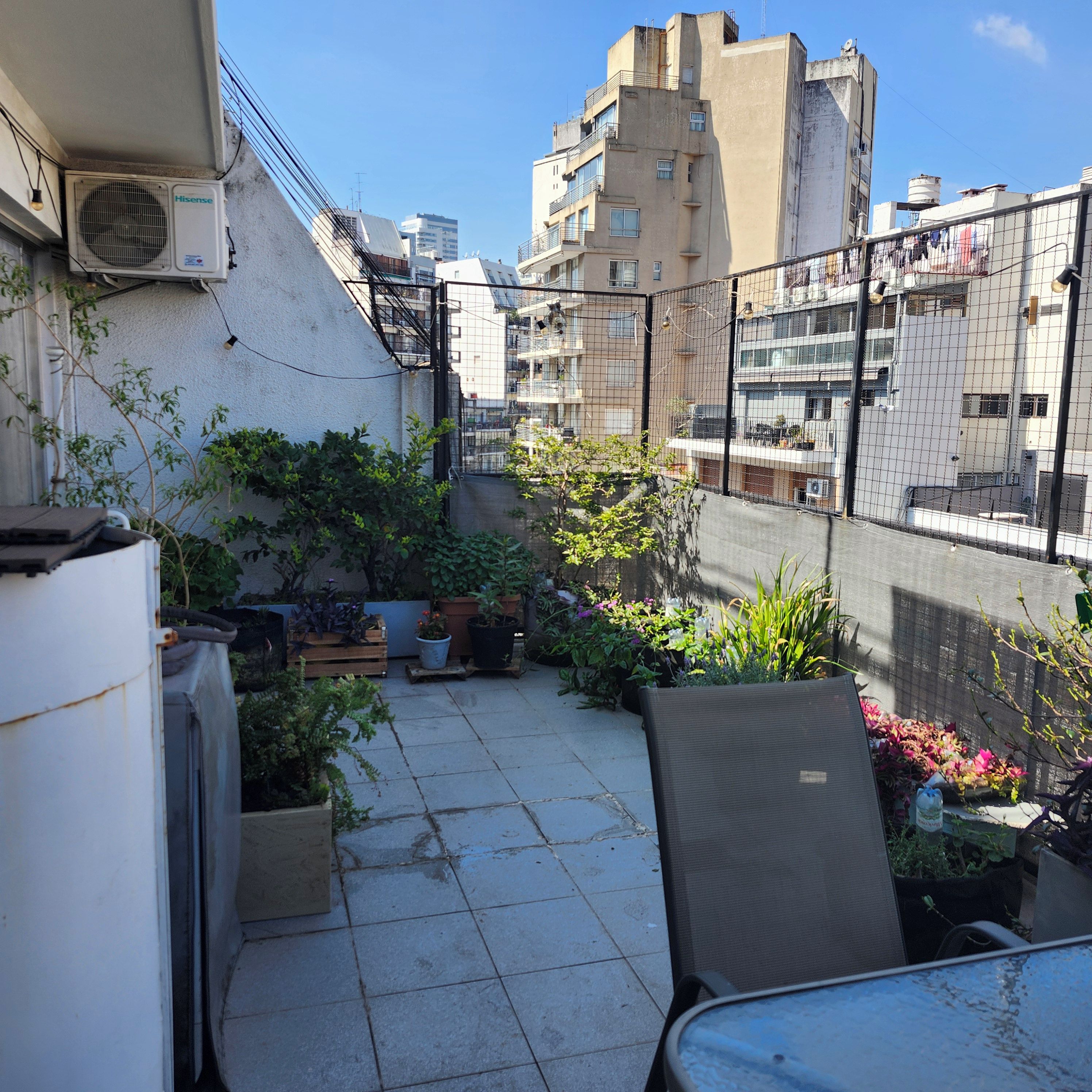 An outdoor patio area with a table and chairs, surrounded by potted plants and a metal fence.