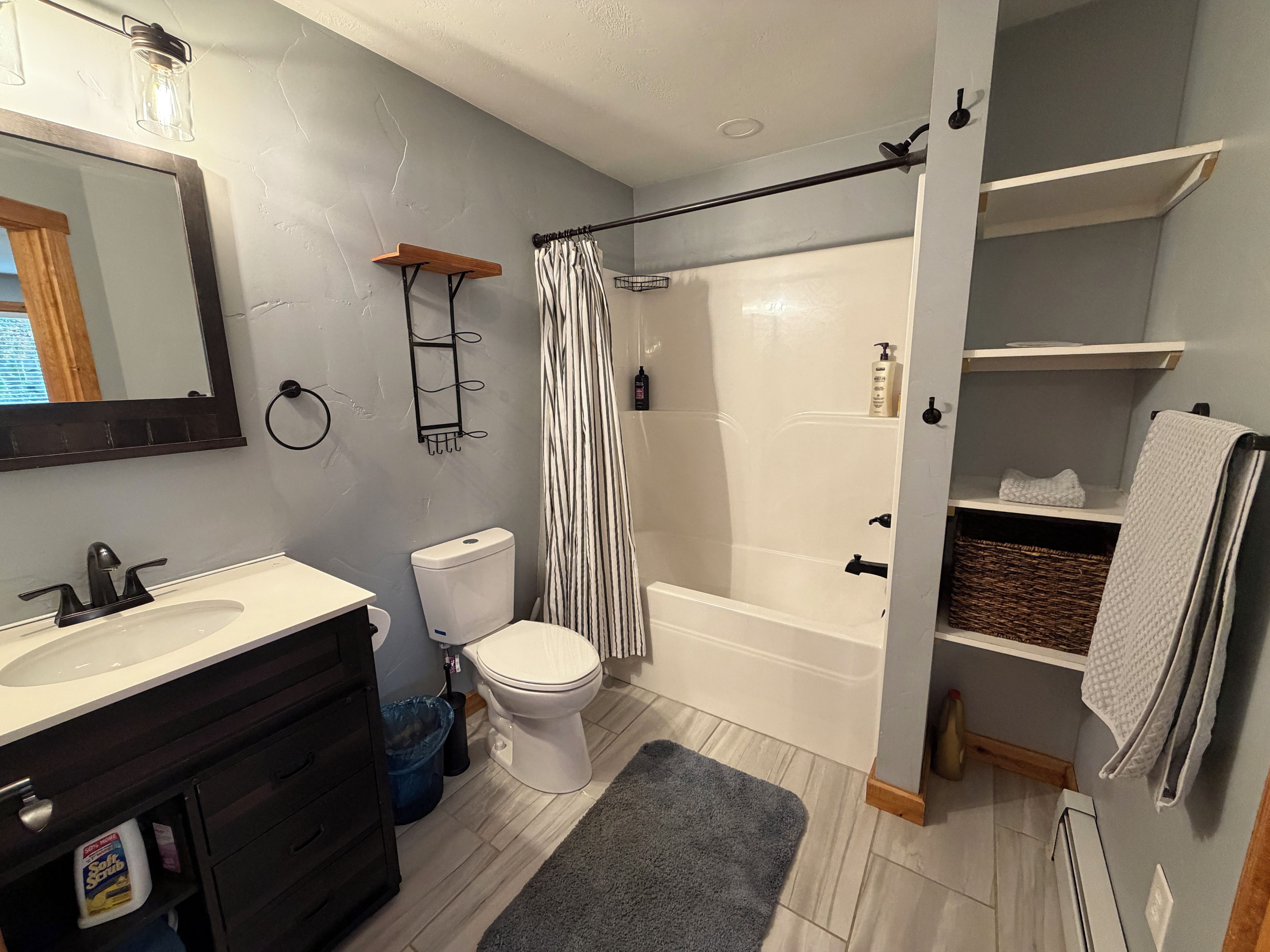 Un baño con lavamanos, inodoro y bañera blancos. Incluye una cortina de ducha, un perchero y un estante con artículos de tocador.