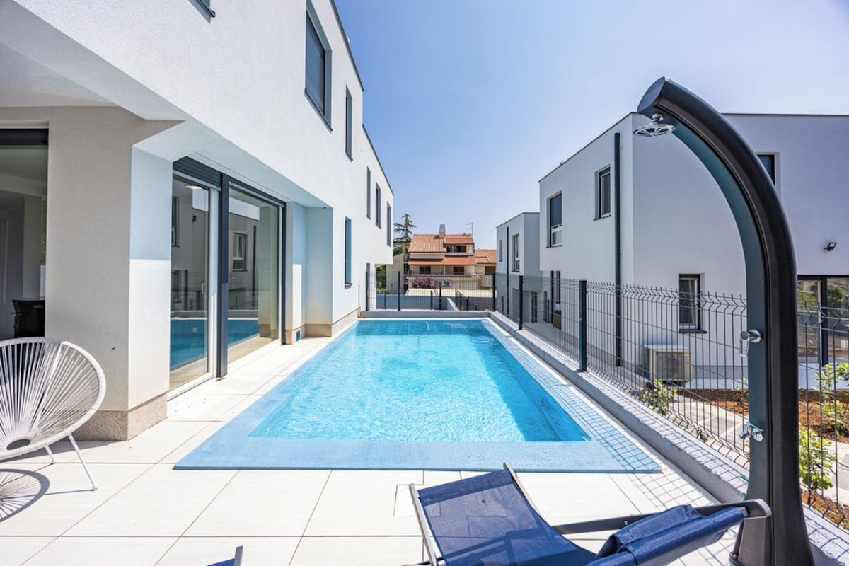 A modern outdoor swimming pool area with a clear blue pool, a lounge chair, and a white chair.