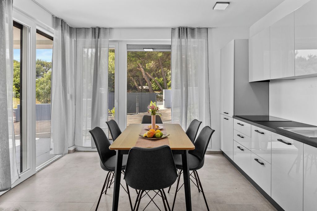 A modern kitchen with a dining table and chairs, a large window with curtains, and a view of greenery outside.