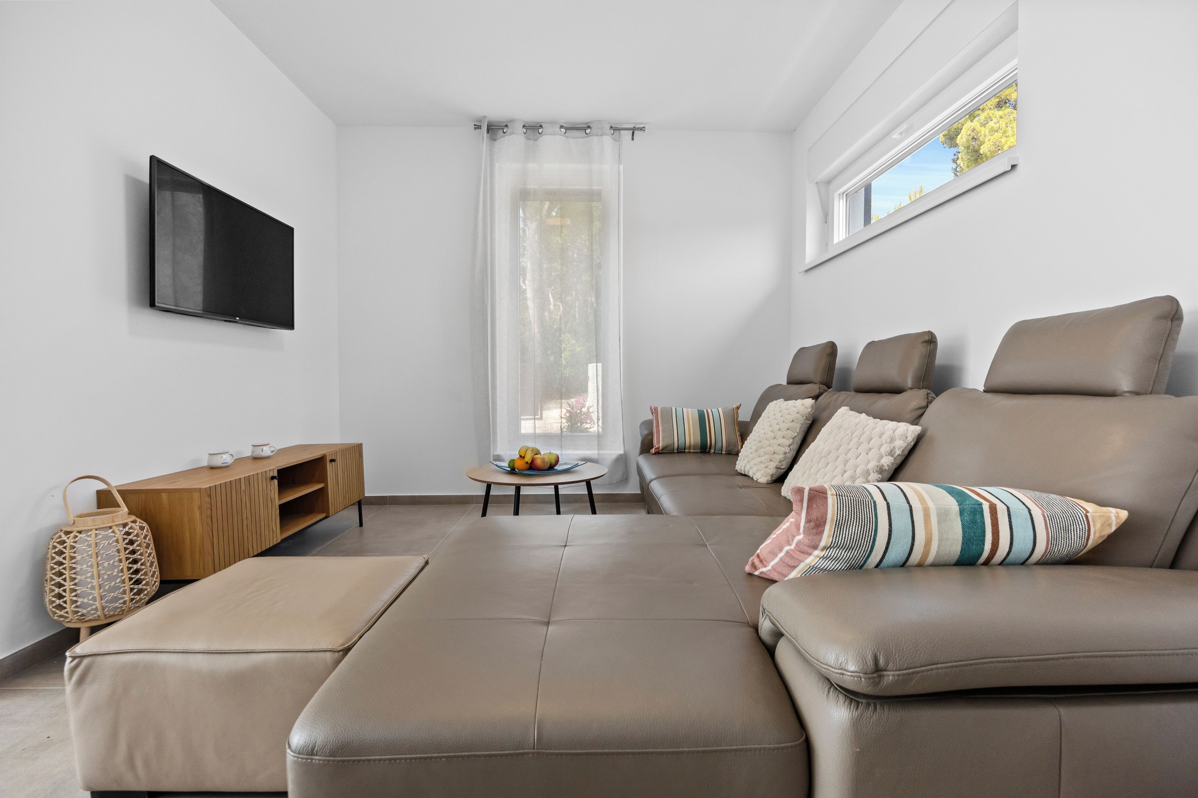 A modern living room with a leather sofa, a wooden side table, a flat-screen TV mounted on the wall, and a large window with a view of trees.