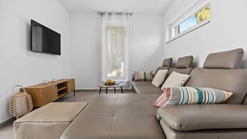 A modern living room with a leather sofa, a wooden side table, a flat-screen TV mounted on the wall, and a large window with a view of trees.