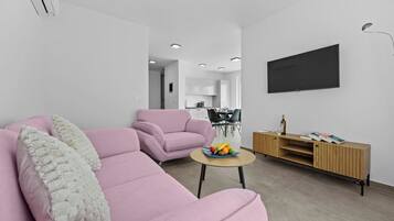 A modern living room with a pink sofa, a small round table with fruit, and a television on the wall.