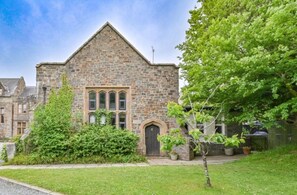 A stone building with a triangular roof, a central entrance with a door, and a small porch area. - Grenville House (Kilkhampton)