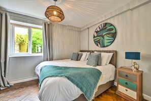 A bedroom with a bed, a bedside table with a lamp, a window with curtains, and a wall art piece. - Grenville House (Kilkhampton)