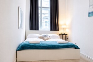 A neatly made bed with a blue bedspread, a bedside lamp, and a view of buildings through a window with dark curtains. - Belvedere on Foot Your Vienna 1BR (Vienna)