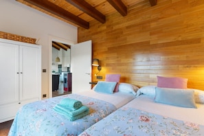 A room with two beds, wooden ceiling beams, a wardrobe, and a kitchenette in the background.
