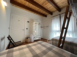 A bedroom with a checkered bedspread, a white wardrobe, a wooden ladder, and a window with sheer curtains.