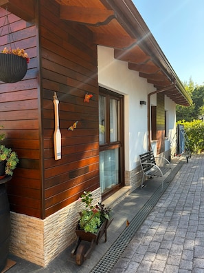A wooden exterior with a stone base, a window, a door, a hanging basket, and a bench.