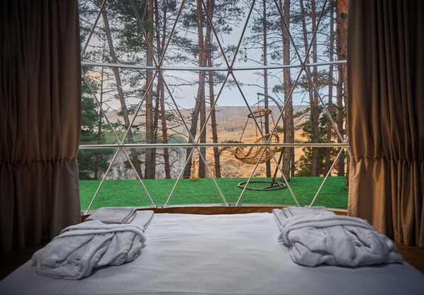 A bed with white linens and towels, a window with a view of a forest and a swing set.