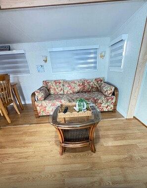 Ein Zimmer mit Holzboden, einem floralen Sofa, einem Korbtisch mit Korb und Fernbedienung sowie einem Fenster mit Jalousien.