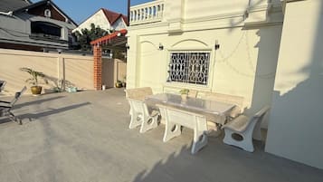 A patio with a table and chairs, a small garden area with potted plants, and a building with a red roof and decorative window grills.