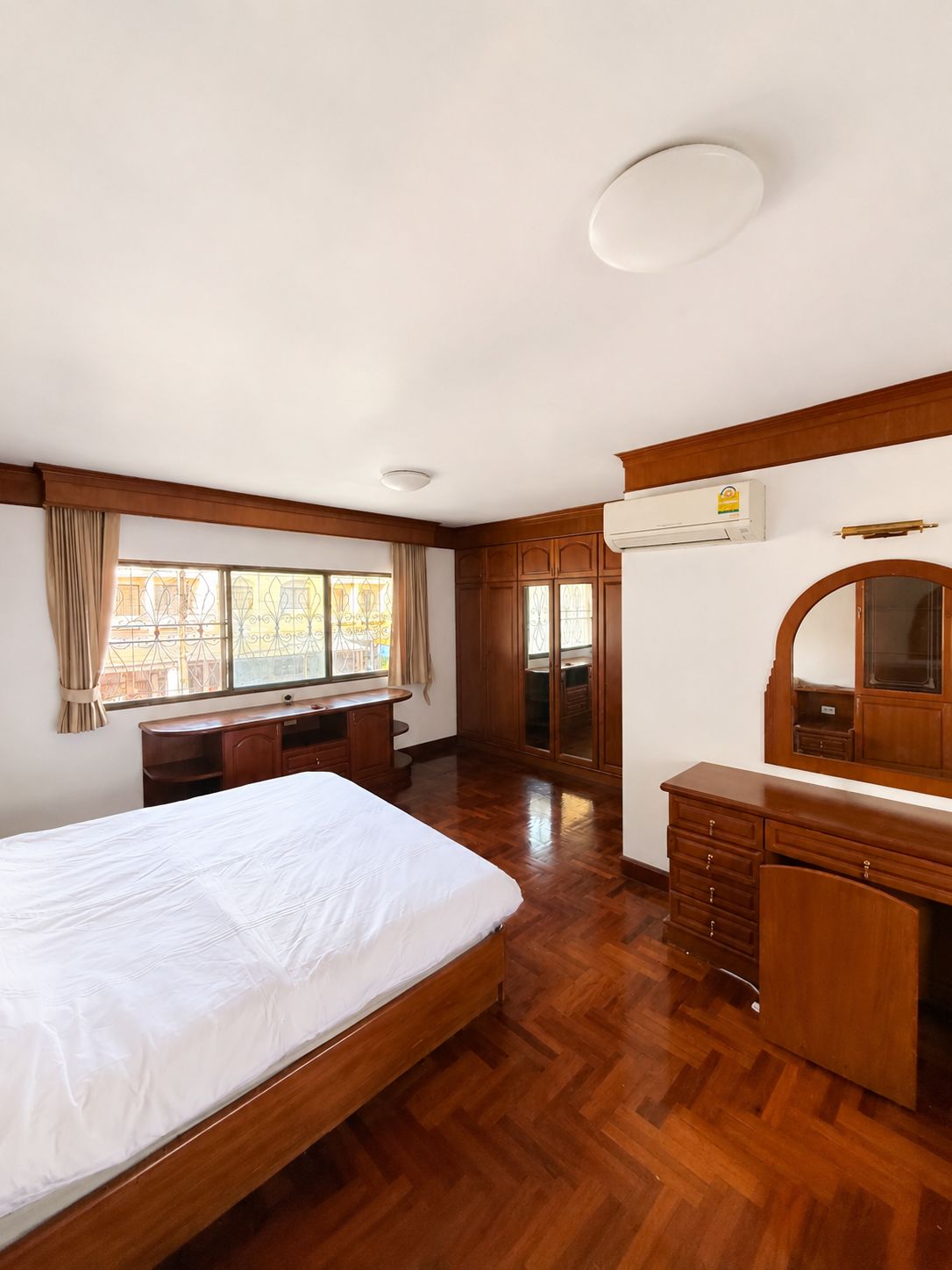 A bedroom with a bed, wooden dresser, vanity, and a window with curtains.