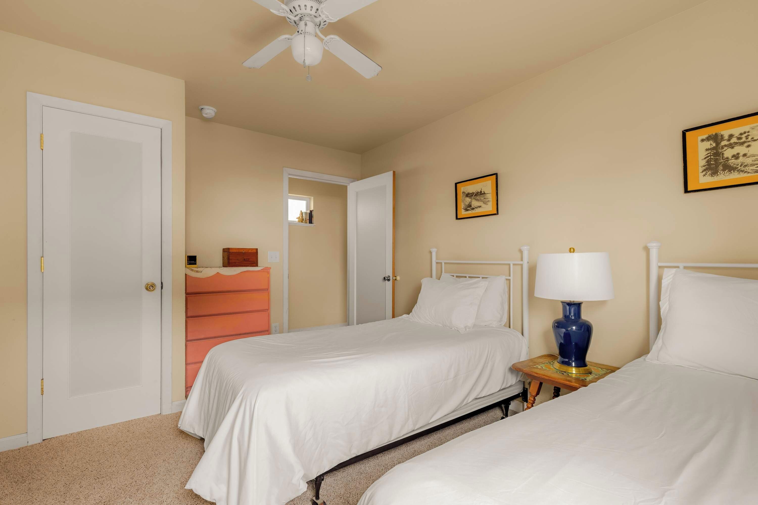A hotel room with two beds, a ceiling fan, a nightstand with a lamp, and a door with a handle.
