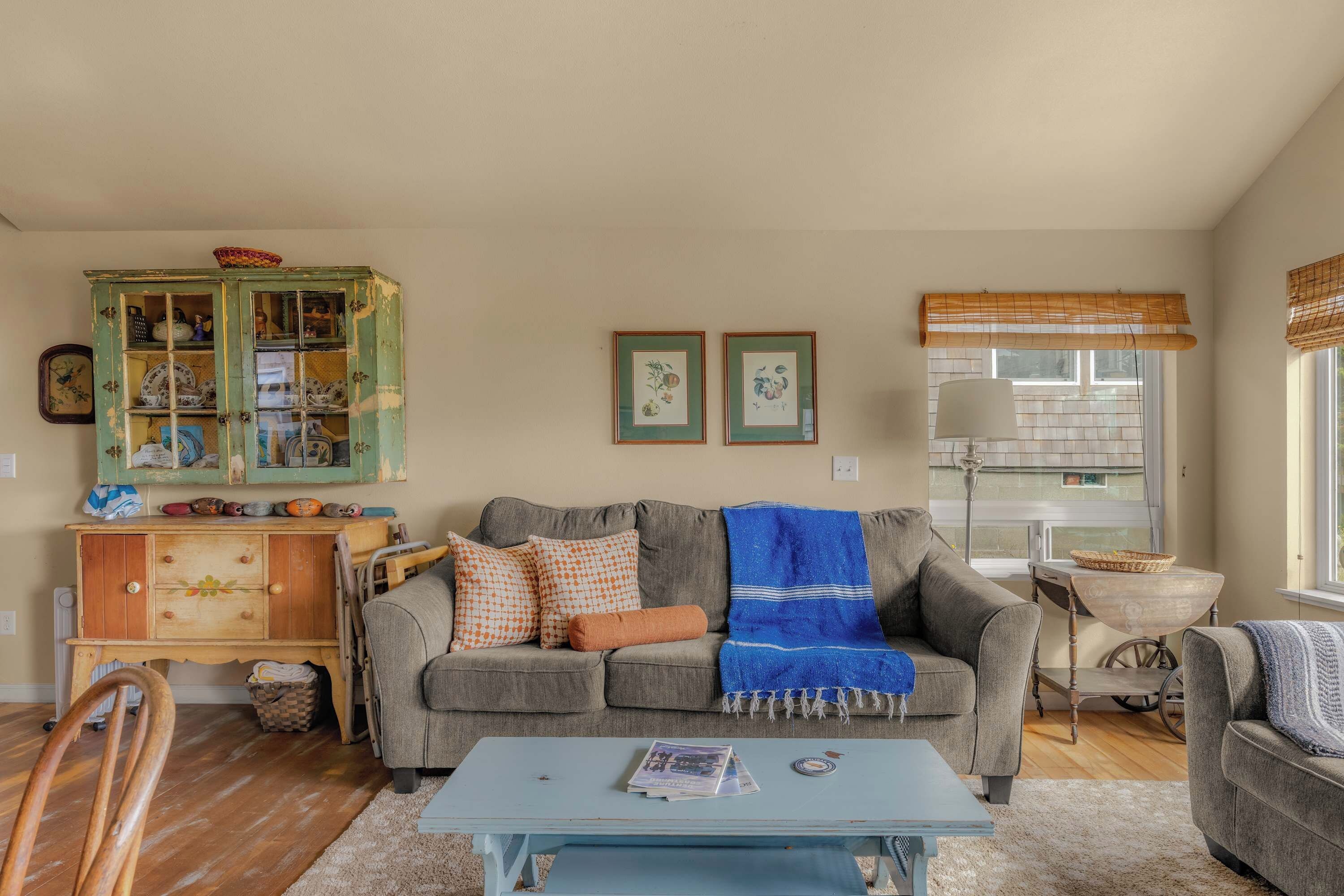A living room with a grey sofa, a blue blanket, a wooden cabinet with glass doors, and a window with a bamboo blind.
